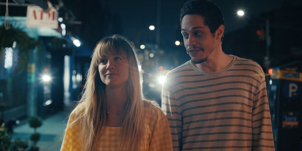 Pete Davidson wears a striped shirt and Kaley Cuoco a yellow checkered dress. They stand next to each other on a dimly lit street.