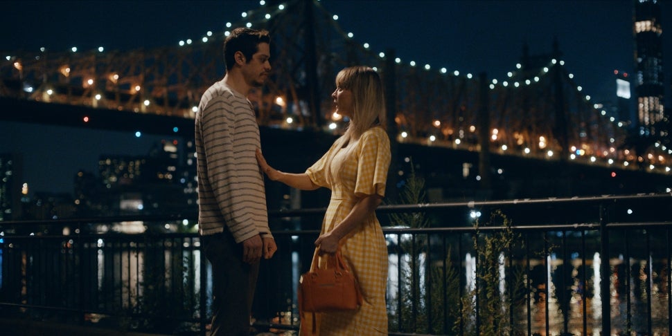 Kaley Cuoco in yellow checkered dress with Pete Davidson in striped shirt. They stand in front of a bridge lit up at night.