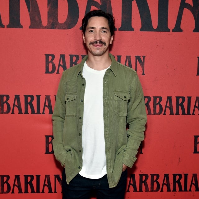 Justin Long in white shirt and green jacket standing in front of red backdrop for 'Barbarian' premier. He has a mustache with dark hair.